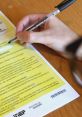 Hand holding a pen writing on a yellow exam sheet with guidelines, highlighting details for the "prova" process.