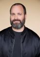 Tom Segura smiling, wearing a black jacket, showcasing his beard and blue eyes against a soft beige background.