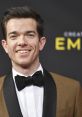 John Mulaney smiles at the Creative Arts Emmys, showcasing his stylish brown suit and bow tie on the red carpet.