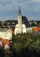 Historic church with a tall spire, surrounded by red rooftops and greenery in a picturesque skyline. Lubor Mrázek's work.