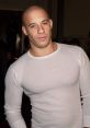 Vin Diesel posing confidently in a fitted white long-sleeve shirt, showcasing his muscular physique and charismatic smile.