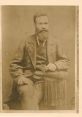 Portrait of a bearded man in 19th-century attire, possibly Hugh Mckinley, seated with decorative fringed cushion.