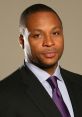 Professional headshot of Gus Johnson, showcasing his polished look with a black suit and purple tie against a neutral background.