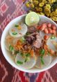 A bowl of soto with tender beef, radish, crispy shallots, green onions, and lime on a vibrant patterned tablecloth.