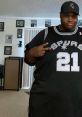 Man in a black Spurs jersey poses with peace sign, showcasing sports fandom in a cozy indoor setting.