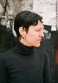 Portrait of a woman in a black turtleneck, styled hair, wearing a gold earring, against a textured background.