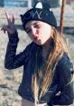 Young girl in shiny black outfit and cat beanie poses playfully, showing peace sign and playful expression against outdoor backdrop.