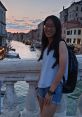 Nicole Kan smiling by a canal at sunset in Venice, wearing a white top and denim shorts, with a backpack.