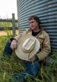 Cory Lunny casually rests by a weathered silo, holding his cowboy hat, surrounded by lush grass and scenic rural vibes.