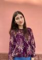 Young woman with long brown hair, wearing a floral blouse against a pink wall, exudes a warm and inviting smile.