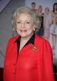 Betty White smiles warmly at a red carpet event, wearing a vibrant red blouse and a decorative brooch.