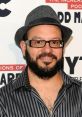 David Cross wearing a stylish hat and glasses, smiling at a promotional event backdrop with bold text.