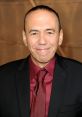 Gilbert Gottfried smiling in a black suit and burgundy shirt at a public event, showcasing his signature comedic style.