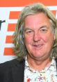 James May smiles at an event, showcasing his distinctive long hair and colorful floral shirt against a vibrant backdrop.