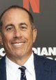 Jerry Seinfeld smiling at a Netflix event, showcasing his signature style and charisma in a blue blazer and striped tie.