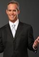 Joe Buck in a dark suit and light tie, smiling confidently with open hands, showcasing his charismatic personality.