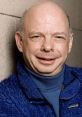 Wallace Shawn smiling, wearing a blue jacket, known for voicing Rex in Toy Story and his work in theater and film.