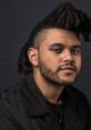 The Weeknd with distinctive hairstyle, wearing black jacket, showcasing his unique look against a dark background.