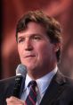 Tucker Carlson speaking into a microphone at a public event, wearing a suit and a tie, with a focused expression.