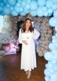 Yimi Charry Rios glowing in a white dress at a festive balloon-decorated event, showcasing her blossoming pregnancy.