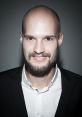 Portrait of Daniel Cz with a shaved head and beard, smiling in a smart black suit and white shirt against a dark background.