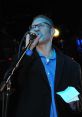 Alex Gutman performing passionately on stage, holding a microphone and a paper, wearing a stylish black coat.