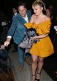 Florence Zack in a vibrant yellow dress, being assisted by a gentleman, exiting a venue during a nighttime event.