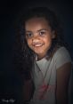 Smiling girl with curly hair wearing a ballet-themed shirt, showcasing a warm expression against a dark background.