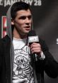 Dominick speaking at a UFC event, wearing a hoodie and a graphic t-shirt, holding a microphone, engaging the audience.