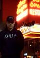 Person wearing a "Chills" hoodie and cap stands in front of a vibrant neon restaurant sign at night, exuding a mysterious vibe.