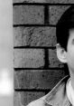 Close-up of a young man with dark hair and a light suit, set against a textured brick wall, evoking a thoughtful mood.