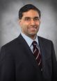 Portrait of Mahish Bhatia in a suit, smiling confidently against a gray backdrop, embodying professionalism and approachability.