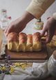 Freshly baked bread being handled with care on a wooden board, surrounded by flowers and rustic kitchen items.