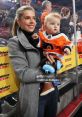 Cheerful woman holds baby wearing Flyers jersey, both enjoying a hockey game with ear protection for the little one.