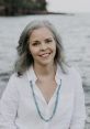 Smiling woman with gray hair, wearing a white shirt and turquoise necklace, by a serene water backdrop.