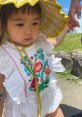 Adorable child in embroidered white dress and yellow sun hat, enjoying a sunny day outdoors with a loved one.