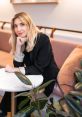 Natalia Albin seated at a table, showcasing a stylish outfit, surrounded by indoor plants in a modern cafe setting.