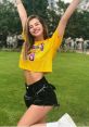 Cheerful woman in a yellow crop top and shorts poses joyfully in a green park, showcasing vibrant summer fashion and confidence.