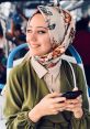 Smiling young woman in a floral scarf and green outfit, using her phone on public transport. Asya Demirci Kargılı style.