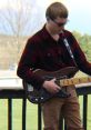 Jalyn Groff performing outdoors with an electric guitar, wearing a black and red plaid shirt and sunglasses.