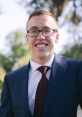 Ethan Ream smiling confidently in a navy suit with a burgundy tie against a lush outdoor background.