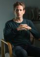 Dennis Reynolds from It's Always Sunny in Philadelphia, sitting thoughtfully in a chair, hands clasped, wearing a dark shirt.