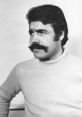 Portrait of Alain Antelme, showcasing a confident expression with a distinctive mustache and a light sweater.