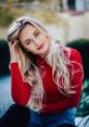 Smiling young woman with long blonde hair, wearing a red sweater, poses outdoors with a relaxed and inviting expression.