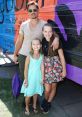 Julia Fachinelli poses with two girls in colorful outfits by a vibrant vehicle, promoting joy and community engagement.