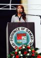 Samar Neweshy delivers a speech at an event, with the Province of Samar seal and floral arrangements in the background.