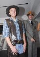 Two men in costumes: one in a cowboy outfit, the other in vintage formal attire, smiling at a lively event.