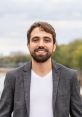 Guilherme Silveira smiling outdoors, wearing a gray blazer and white shirt, with a blurred natural background.