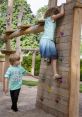 Children playing at a playground, one climbing a wooden wall while the other encourages them. Fun outdoor activity for kids.