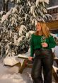 A woman in a green sweater and black pants holds a glass of wine, surrounded by snow-covered trees.